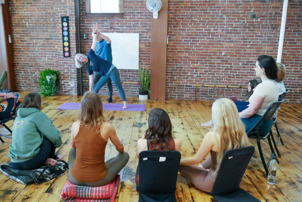 a circle of seated students in a yoga studio watching a man assist a woman with a side angle yoga post